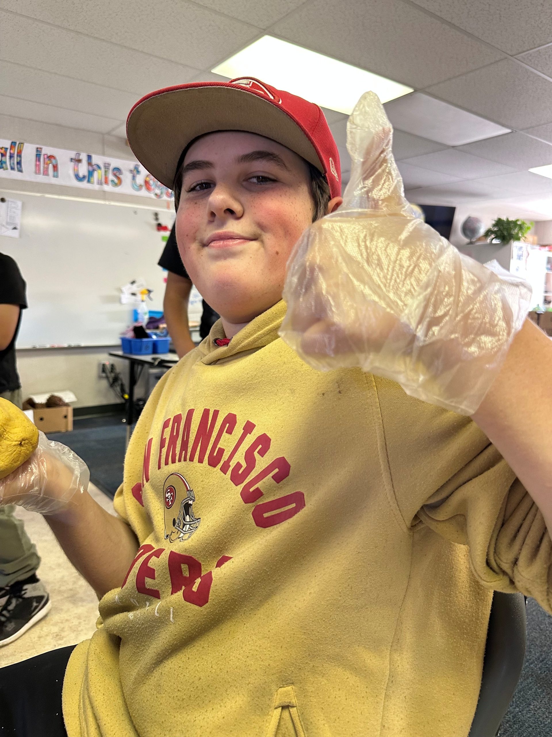Noah Johnson – Student of the Month Noah Johnson gives a thumbs up while smiling in class, wearing a 49ers hoodie and plastic gloves.