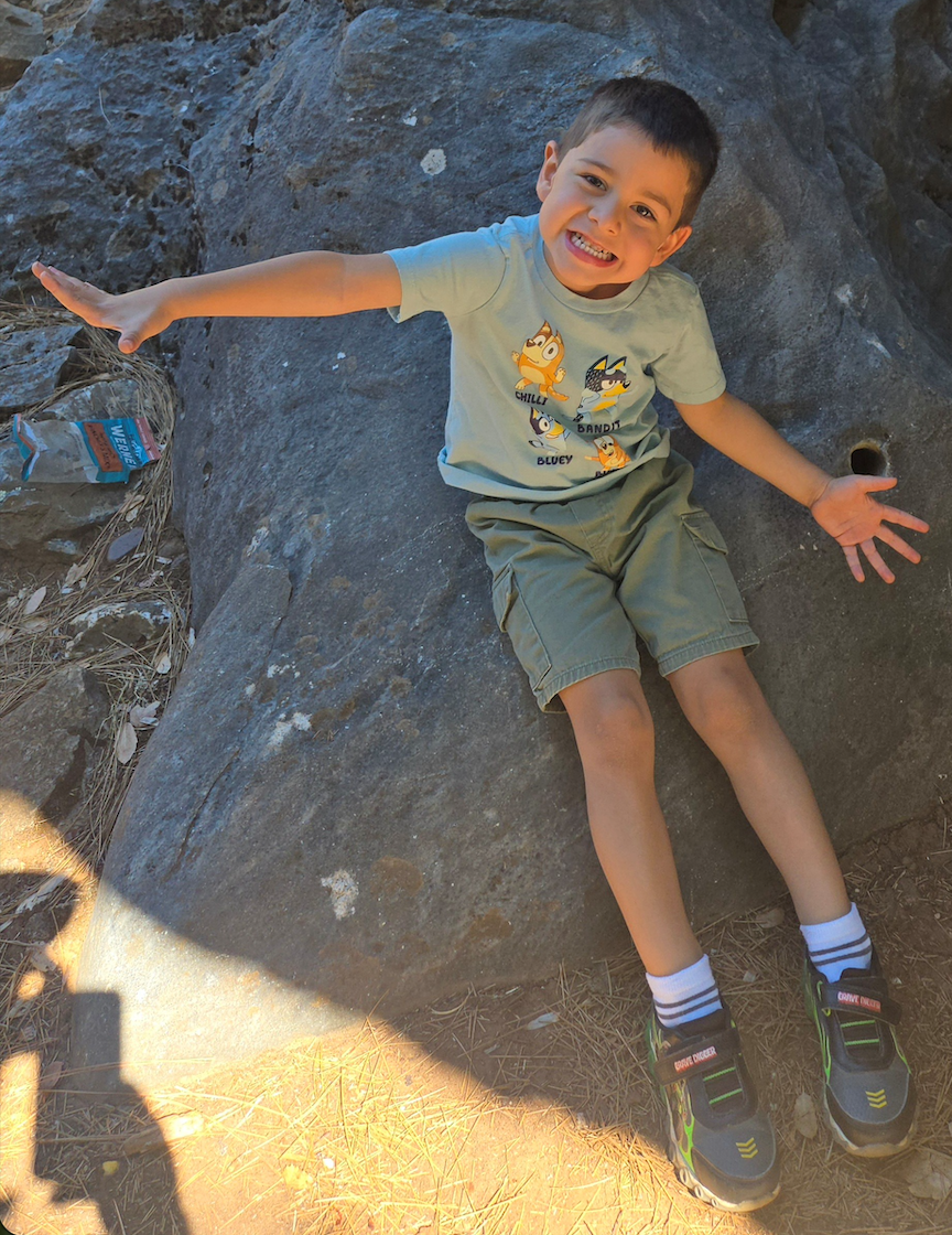 Logan smiles while sitting on a large rock outdoors, arms outstretched in a playful pose.